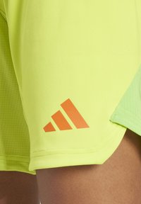 Neon gele sportshorts met een gestructureerd oppervlak en een oranje Adidas-logo op de onderste zoom, met een contrasterend groen zijkantpaneel.