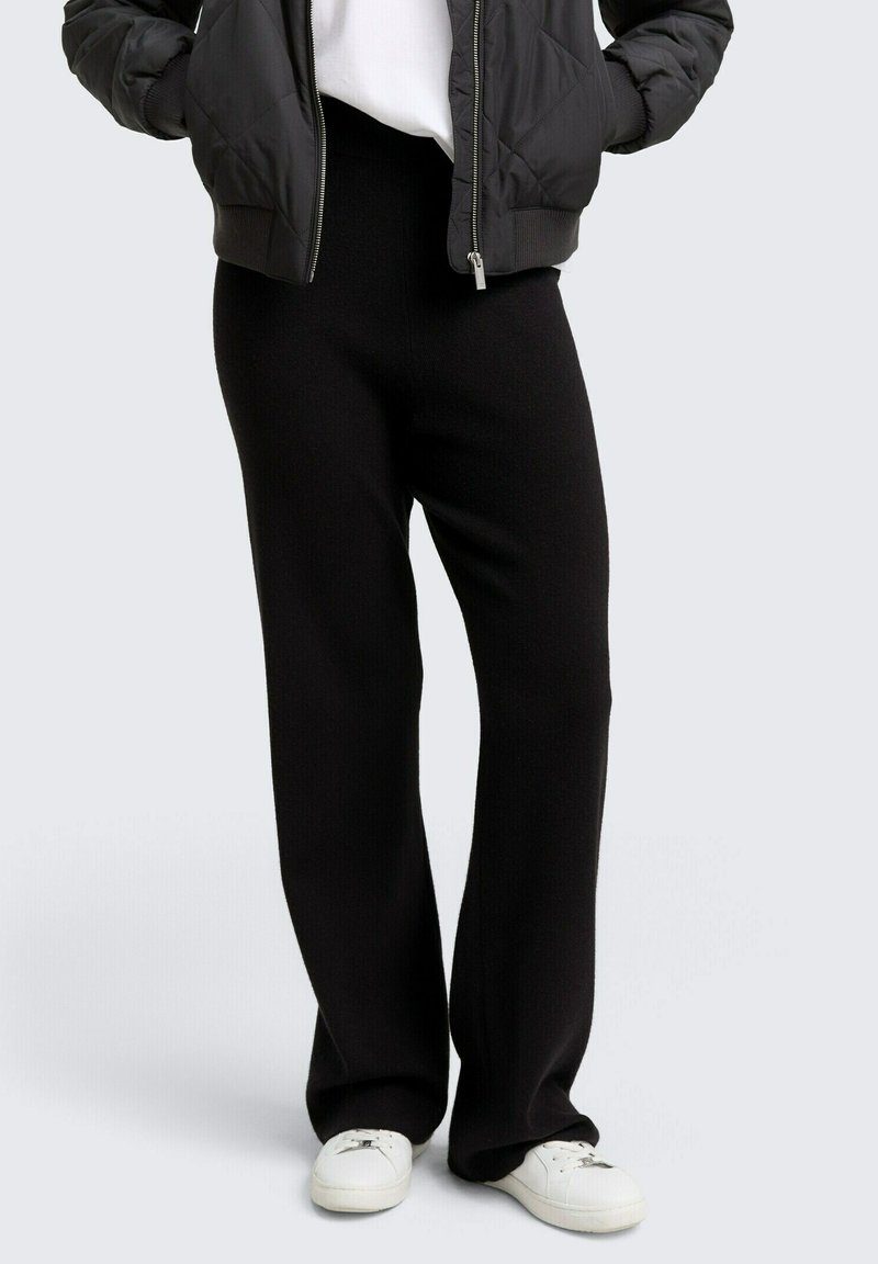 Persona che indossa pantaloni neri a gamba larga, sneakers bianche, una maglietta bianca e un giubbotto imbottito nero con le mani in tasca.