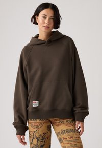 Felpa beige oversize con tasca a marsupio e orlo a costine. Il tessuto appare morbido e testurizzato, con un'etichetta sul davanti.