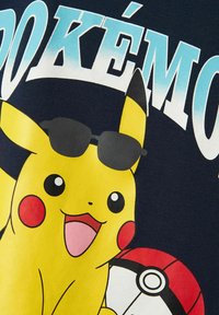 Pikachu amarillo con gafas de sol negras, en una camiseta azul marino. Presenta una Poké Ball y el texto "POKÉMON" en azul claro encima del personaje.