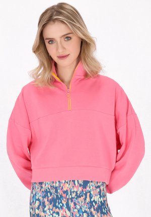 Junge Frau mit blondem Haar, die einen knallpinken Reißverschluss-Sweatshirt und bunte gemusterte Leggings trägt, steht vor einem weißen Hintergrund.