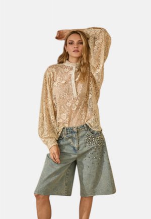 Donna che indossa una blusa di pizzo floreale beige con maniche lunghe e corti di denim blu larghi con ornamenti di strass.