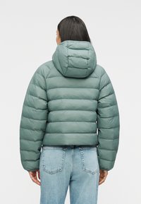 Μπουφάν με επένδυση σε απαλό πράσινο χρώμα με κουκούλα. Διαθέτει κουτί σχήμα και καπιτονέ υφή. Συνδυάζεται με ανοιχτόχρωμο τζιν παντελόνι.