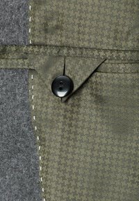 Grün gemusterter Stoff mit einer strukturierten Oberfläche, mit einem schwarzen Knopf und grauem Material auf einer Seite. Genähte Details in weißem Faden.