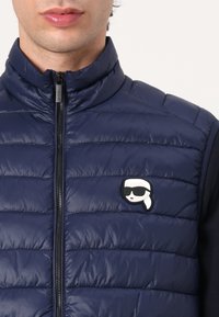 Marineblauwe puffer vest met een hoge kraag, geometrisch patroon en een rits aan de voorkant. Bevat een opvallend wit en zwart logo op de borst.