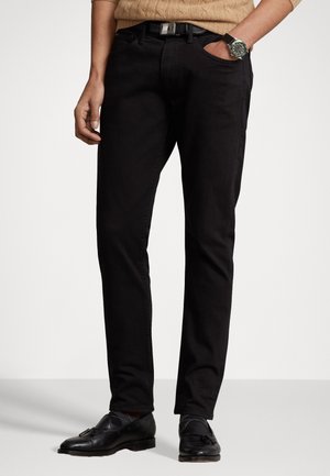 Homme portant un pantalon noir slim, des mocassins noirs à pampilles, un pull beige en maille torsadée, une ceinture noire et une montre noire.