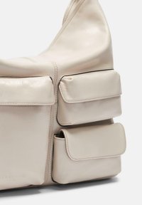 Beżowa skórzana torebka z trzema przednimi kieszeniami z klapką, widocznym szyciem i lekko opadającym kształtem na białym tle.