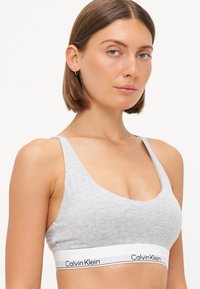 Grijze katoenen bralette met een ronde hals, brede bandjes en een witte band met het Calvin Klein-logo in het zwart. Soepele textuur, minimalistisch ontwerp.