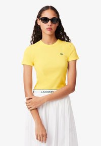 Geel geribbeld T-shirt met korte mouwen, voorzien van een klein groen logo. Gecombineerd met een witte rok, die een gestructureerd ontwerp en een elastische tailleband heeft.