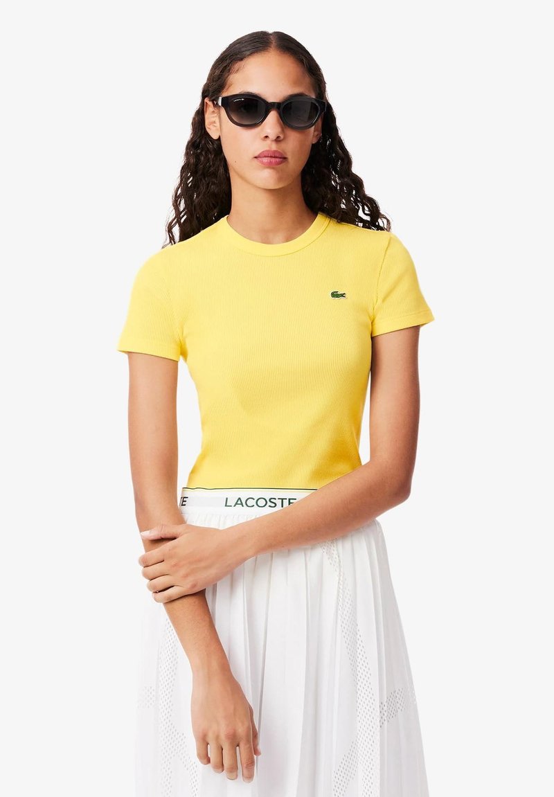 Geel geribbeld T-shirt met korte mouwen, voorzien van een klein groen logo. Gecombineerd met een witte rok, die een gestructureerd ontwerp en een elastische tailleband heeft.