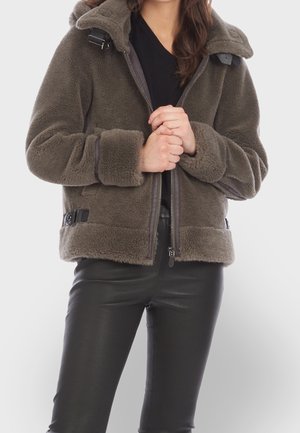 Mujer con chaqueta de forro polar marrón con detalles de hebilla, cerrada hasta la mitad, combinada con pantalones de cuero negros, con las manos entrelazadas a la altura del abdomen.