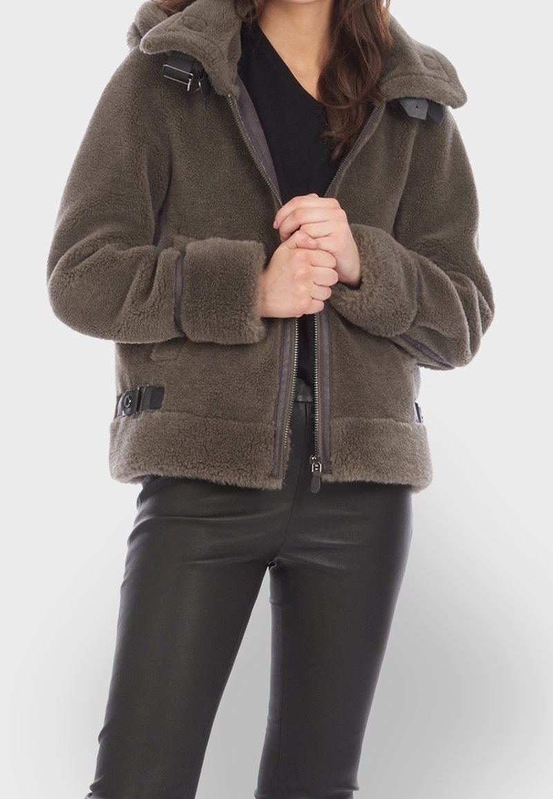 Femme portant une veste en polaire marron avec des détails à boucles, zippée à mi-hauteur, associée à un pantalon en cuir noir, mains jointes au niveau de la taille.