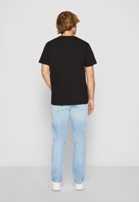 Musta puuvillapaita, jossa on lyhyet hihat, yksinkertainen pyöreä kaula-aukko, yhdistettynä vaaleansinisiin farkkuihin ja valkoisiin lenkkareihin, nähtynä takaa.