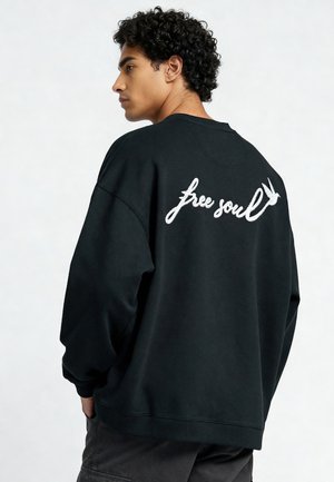 Giovane uomo con capelli ricci, indossa una felpa nera con la scritta bianca "free soul" e un disegno di un uccello sulla schiena, mani in tasca, rivolto verso sinistra.