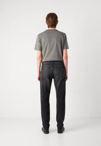Jeune personne de dos, portant un t-shirt gris à manches courtes, un jean noir et des chaussures noires sur un fond clair uni.