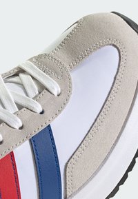 Baskets blanches avec des accents en daim gris et des rayures bleues et rouges. Présente un dessus en tissu lisse, des lacets ronds et une semelle en caoutchouc noir texturé.