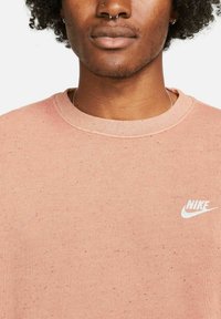 Růžová fleecová mikina s kulatým výstřihem, na levé straně hrudi s bílým logem Nike. Měkká textura se skvrnitým vzorem.