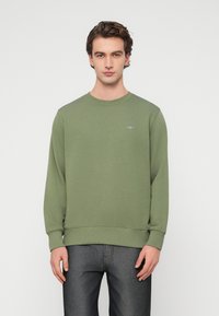 Πράσινο βαμβακερό φούτερ με μακριά μανίκια, στρογγυλή λαιμόκοψη και ριμπ μανσέτες. Φέρει ένα μικρό λογότυπο στο στήθος. Συνδυάζεται με σκουρόχρωμο γκρι τζιν.