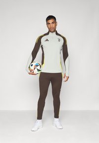 Tenue de sport comprenant un haut zippé gris clair avec des accents marron et jaune et un legging marron assorti, tenant un ballon de football.
