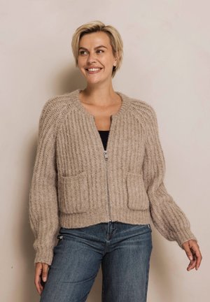 Beige gestrickte Strickjacke mit Reißverschluss vorne, mit geripptem Muster, zwei Taschen vorne und langen Ärmeln. Kombiniert mit blauer Jeans.