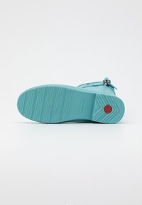 Une sandale en caoutchouc turquoise présente une semelle texturée avec des motifs horizontaux et en zigzag, une boucle sur le côté et un logo d'accent rouge.