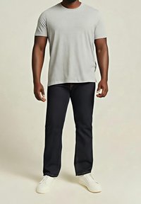 Homme portant un T-shirt gris clair à manches courtes, un pantalon foncé et des chaussures blanches sans lacets, debout devant un fond neutre.