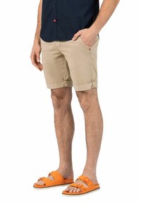 Beige Baumwollshorts mit umgeschlagenen Säumen und seitlichen Taschen, kombiniert mit orangen Gummisandalen mit zwei verstellbaren Riemen.