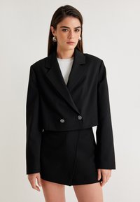 Blazer nero cropped con chiusura a doppio petto, maniche lunghe, vestibilità sartoriale, abbinato a una gonna mini coordinata. Texture liscia.