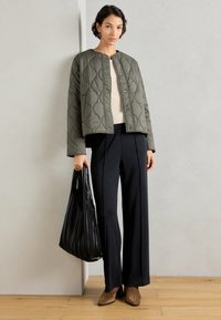 Gequiltede olijfgroene jas met drukknoopsluitingen, gedragen over een crèmekleurige trui, gecombineerd met zwarte wijde broek en een zwarte geplooide tote bag.