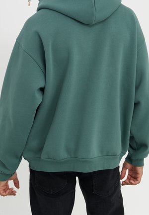 Persoon die een losse groene capuchonsweater en zwarte spijkerbroek draagt, van achteren gezien tegen een effen lichte achtergrond.
