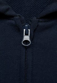 Indumento blu navy con zip e texture a costine. Presenta un tirazip argentato e una sovrapposizione di tessuto liscio nell'area del colletto.