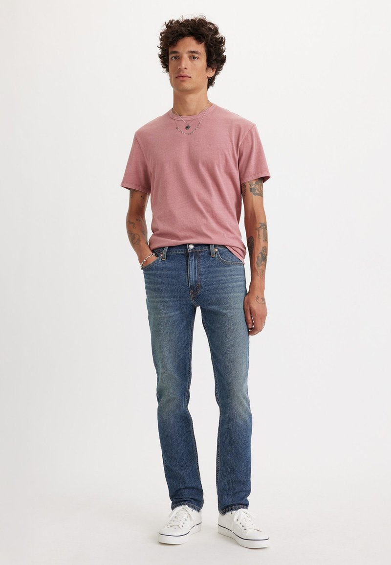 T-shirt in cotone rosa chiaro abbinato a jeans in denim blu. I jeans hanno una vestibilità dritta e una leggera scoloritura; il modello indossa sneaker bianche.