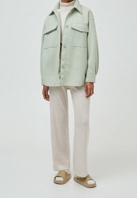 Camicia button-up oversize di colore verde chiaro con due tasche frontali, abbinata a pantaloni a gamba larga ribattuti beige e sandali a scivolo color tan.