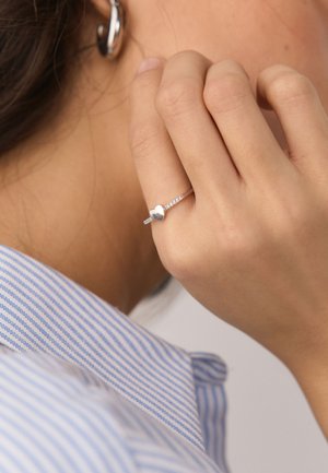 Hand touching ear with silver heart-shaped ring on finger and silver hoop earring, wearing blue and white striped shirt collar.