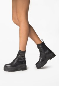 Bottes de combat noires en cuir avec une semelle épaisse, devant à lacets et une fermeture éclair latérale. Elles disposent d'une languette de maintien et d'un motif de semelle texturé.