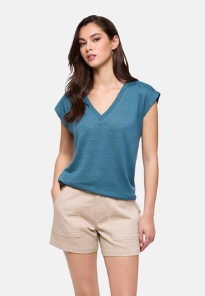 Joven mujer con cabello largo y oscuro que lleva una camiseta azul de cuello en V y pantalones cortos beige claro, de pie con una mano en el bolsillo.