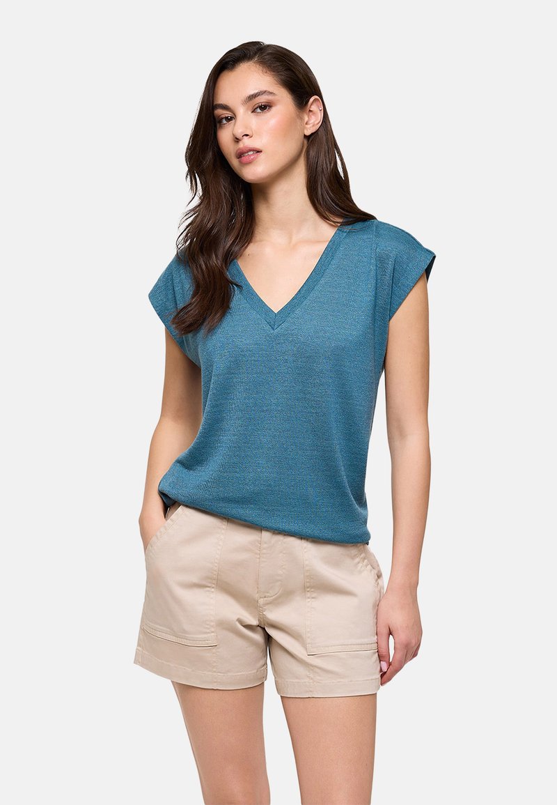 Joven mujer con cabello largo y oscuro que lleva una camiseta azul de cuello en V y pantalones cortos beige claro, de pie con una mano en el bolsillo.