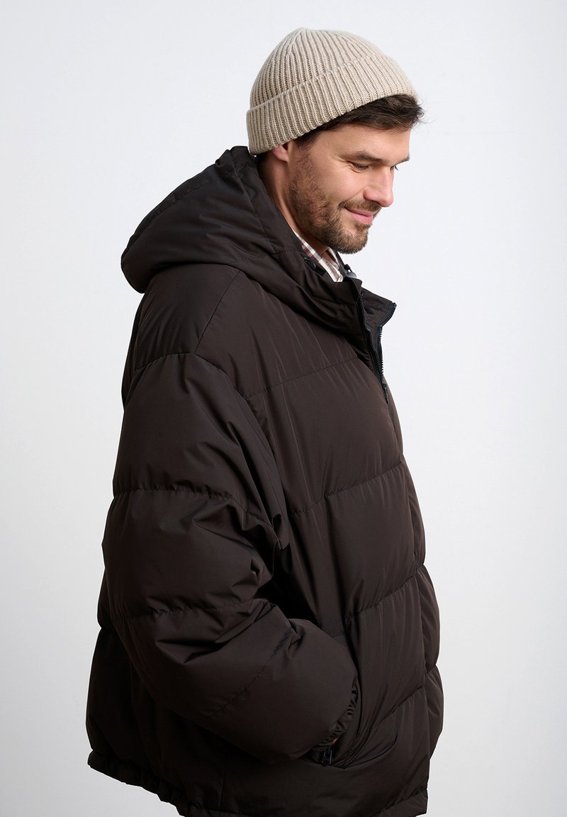 Brązowa pikowana kurtka o fakturowanej powierzchni, o luźnym kroju, z kapturem i bocznymi kieszeniami, zestawiona z beżową dzianinową czapką.