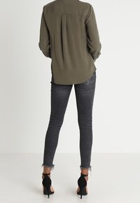 Woman in olive green long-sleeve blouse, dark gray frayed hem skinny jeans, and black high-heeled ankle-strap shoes, standing facing away.