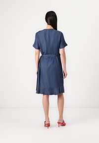 Robe en denim à manches courtes, avec un ourlet évasé et un détail de lien à la taille. La couleur est bleu foncé. Portée avec des chaussures à talons rouges.