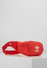 Marsupio rosso in tessuto con due compartimenti con zip, caratterizzato da un logo Adidas bianco e un accento a strisce sulla tracolla regolabile.