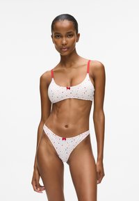 Moteris, vilkinti baltą liemenėlę ir kelnaites su raudonais taškeliais ir mažomis širdelėmis, raudonais dirželiais ir raudonomis kaspinėliais, stovi prieš paprastą baltą foną.