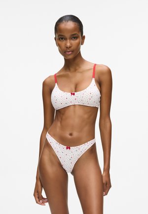 Kvinne iført hvit bh og truser med røde prikkete og små hjerte mønstre, røde stropper og røde sløyfer, som står foran en enkel hvit bakgrunn.