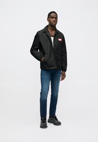 Melna jaka ar pogām un sarkanu logotipa uzliku, valkāta virs baltas krekla. Kombinēta ar zilajām džinsām un melniem sporta apaviem.