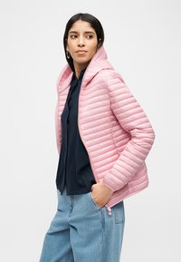 Γυναίκα που φοράει ροζ κουμπωτό, επενδυμένο μπουφάν με κουκούλα πάνω από μαύρο top και μπλε τζιν, με το δεξί της χέρι στην τσέπη, κοιτάζοντας πλάγια.