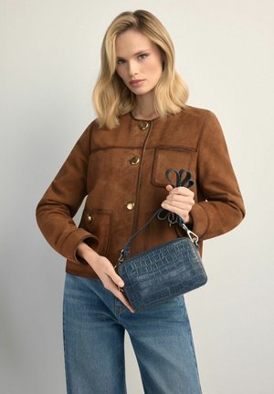 Blondynka w brązowej zamszowej kurtce i niebieskich dżinsach trzyma małą granatową torebkę o fakturze skóry krokodyla za pasek.