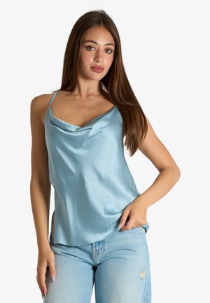 Giovane donna con lunghi capelli castani che indossa un top di raso azzurro chiaro e jeans chiari strappati su uno sfondo uniforme.