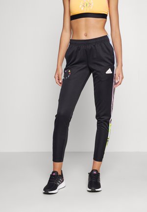 Kobieta w czarnych spodniach dresowych Adidas z kolorowymi napisami i logo, czarnych trampkach oraz krótkim żółtym topie sportowym, stojąca na szarej podłodze.