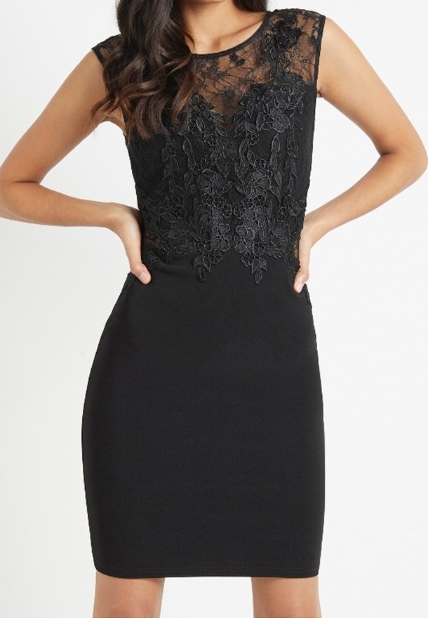 Robe noire sans manches avec jupe cintrée et corsage en dentelle florale transparente, portée par une femme les mains sur les hanches sur fond blanc.