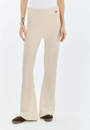 Beżowe, prążkowane spodnie o szerokich nogawkach noszone z brązowymi butami, pokazujące dolną część tułowia i ramiona z tatuażami na białym tle.